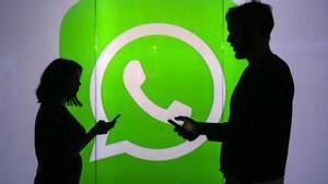 Dos personas hacen uso de sus teléfonos móviles, ante el logotipo de WhatsApp.
